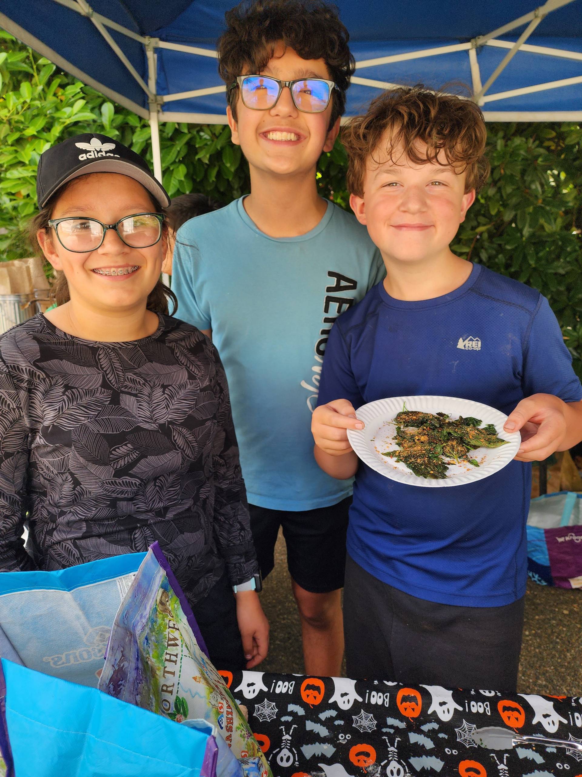 3 campers are standing and smiling.  One is holding a white paper plate with some sauteed stinging nettle on it. 