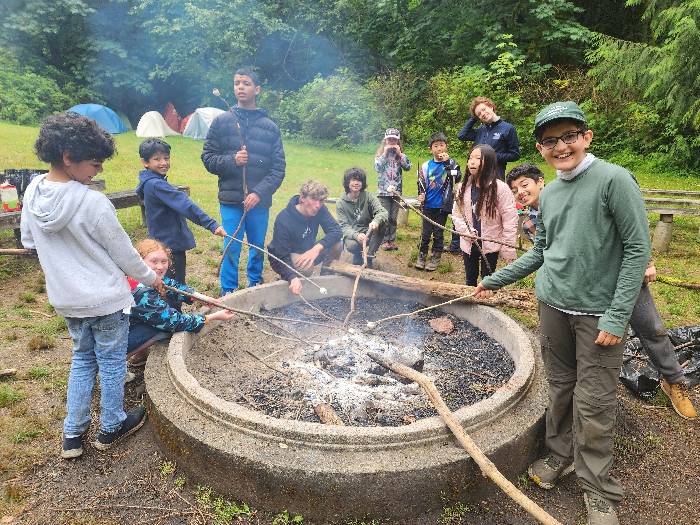 A group of campers is posing around a large cement fire ring.  They are holding various lengths of sticks with marshmallows on the ends.  They are roasting the marshmallows over the embers/coals.  In the background distance there are some tents and a lot of vegetation.