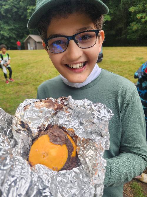 Close-up of a smiling camper who is holding a halved orange filled with chocolate cake on a rumpled piece of aluminum foil.  The orange cake was cooked in the coals of a campfire.