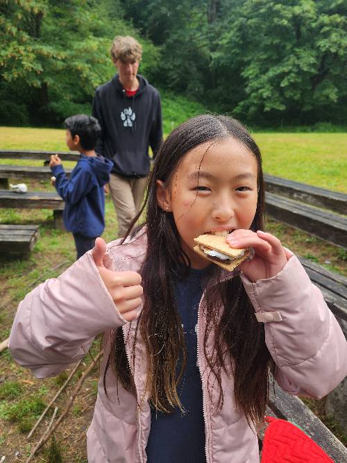 A camper is biting into a s'more while giving a thumbs up with the other hand.  There are 2 campers and some wood benches in the background.