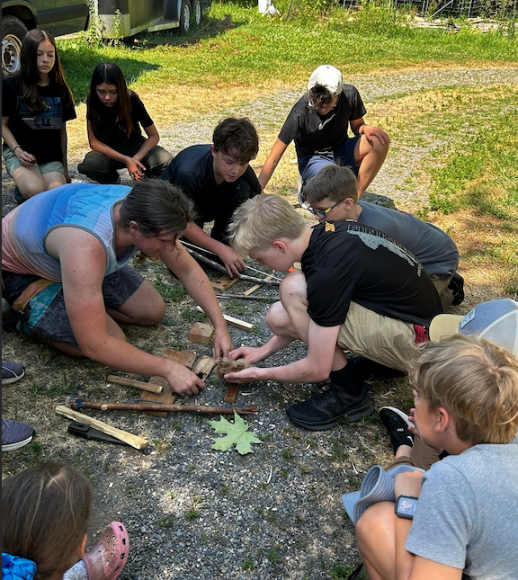 A group of campers is sitting on the ground and looking on as the instructor and assistant put a coal from a bow drill fire-by-friction demonstration into a tinder bundle made of fluffed cedar bark and rafia.