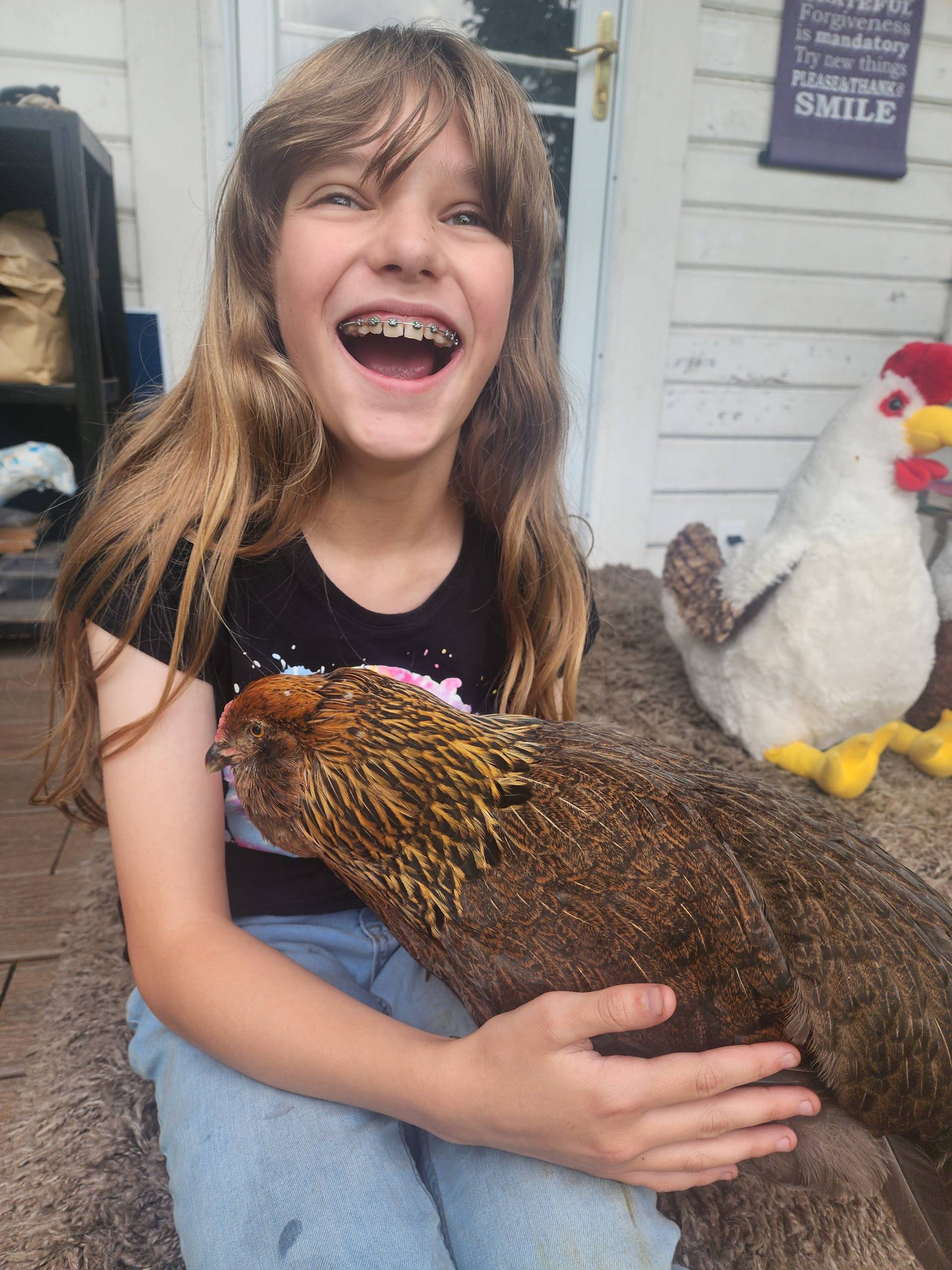 Young girl with a huge smile holding a chicken for the first time.  The chicken is a brown Easter-egger and is on her lap.