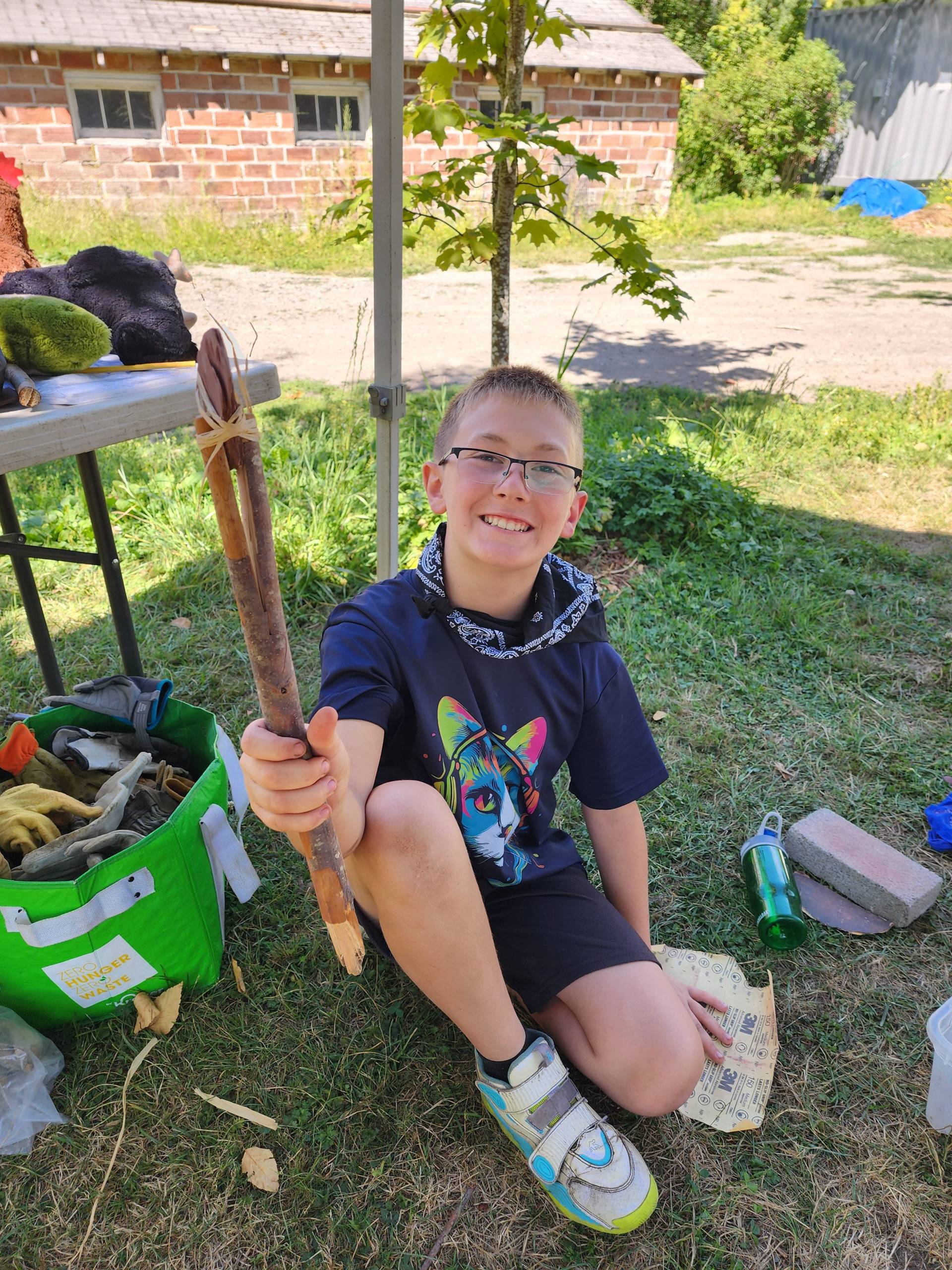 A camper is seated on the ground and smiling up while proudly holding a stone tool that he made using a stick, a smoothed and sharpened rock and raffia. There is a folding table behind him and a green bag next to him that both are holding tools including work gloves and safety goggles.